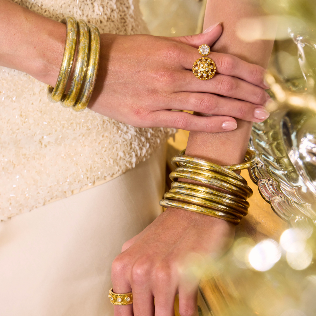 Close up of a woman wearing stacked gold bangle bracelets on both wrists with ornate gold rings and a textured cream outfit beside reflective glassware