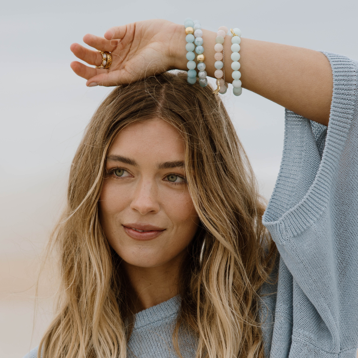 Woman with long blonde hair wearing a light blue sweater and multiple bracelets on a neutral background