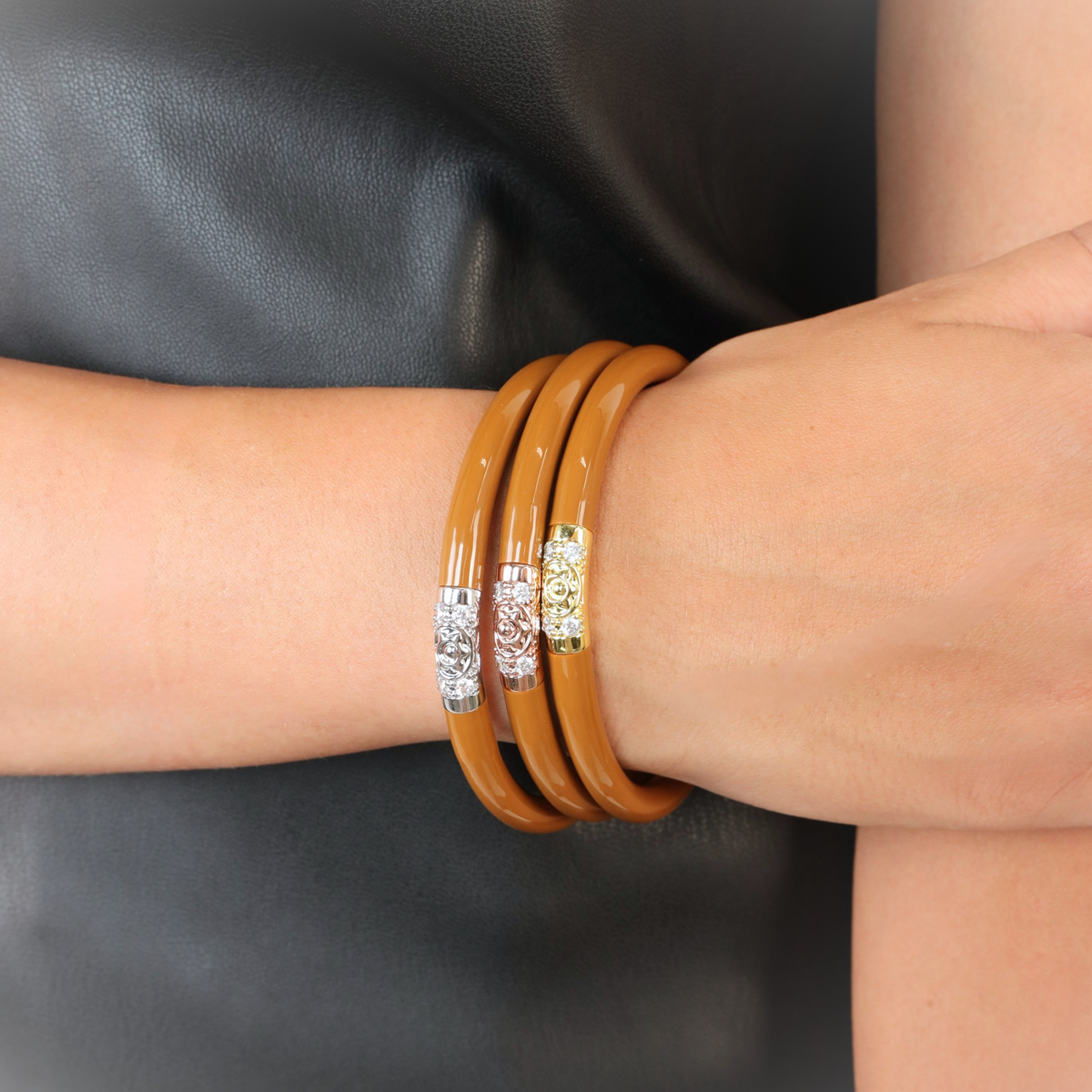Three stacked bangle bracelets on a person's wrist, featuring a matte tan finish with silver and gold-toned filigree center bands, set against a dark leather background.