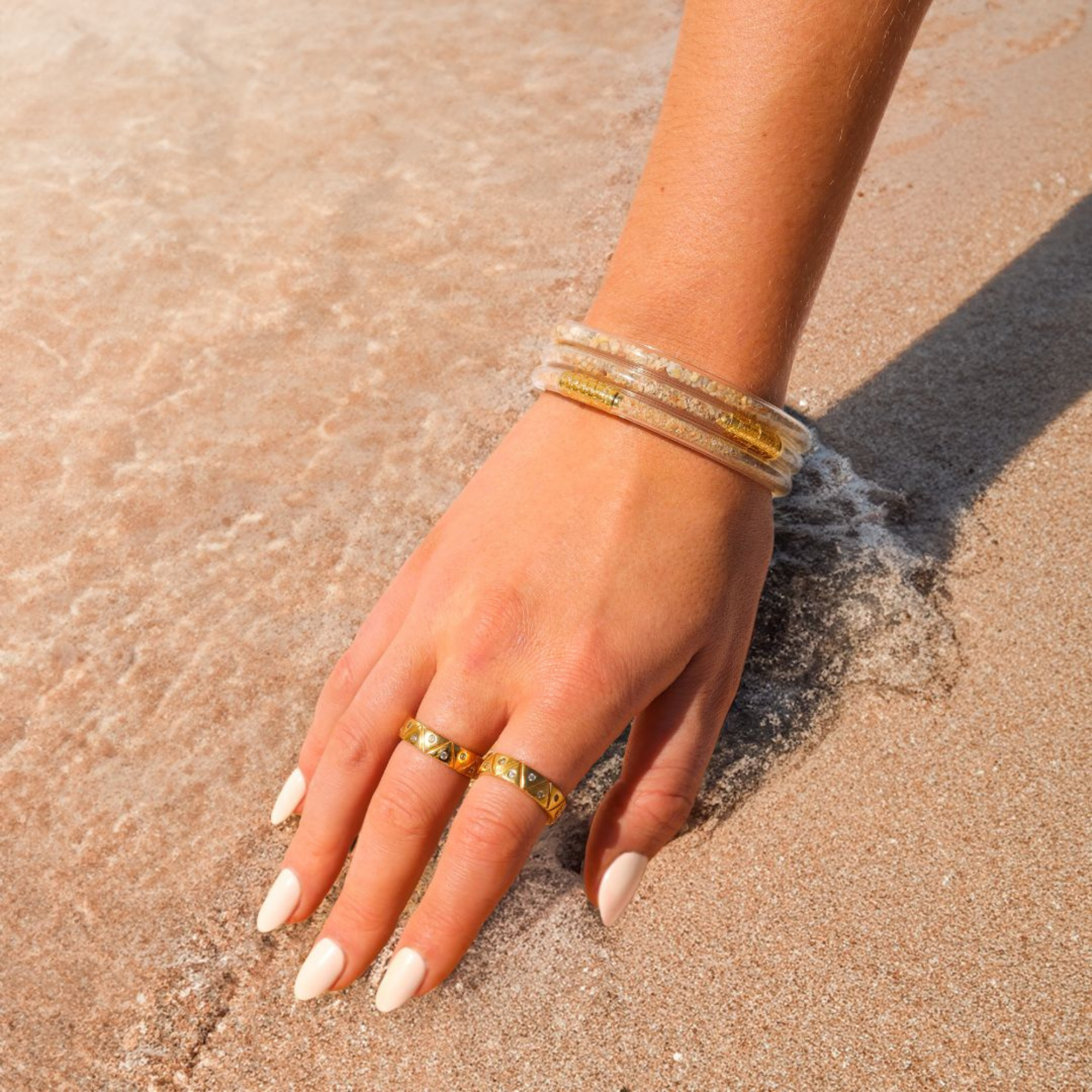 a hand with gold bracelets and rings on sand