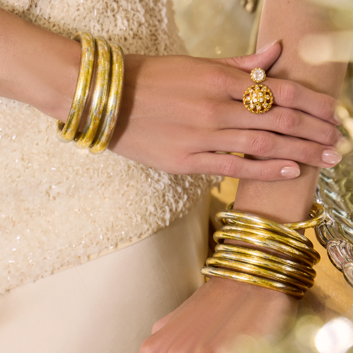 BuDhaGirl gold bangle bracelets for women stacked on both wrists with a matching statement ring against a shimmering neutral backdrop