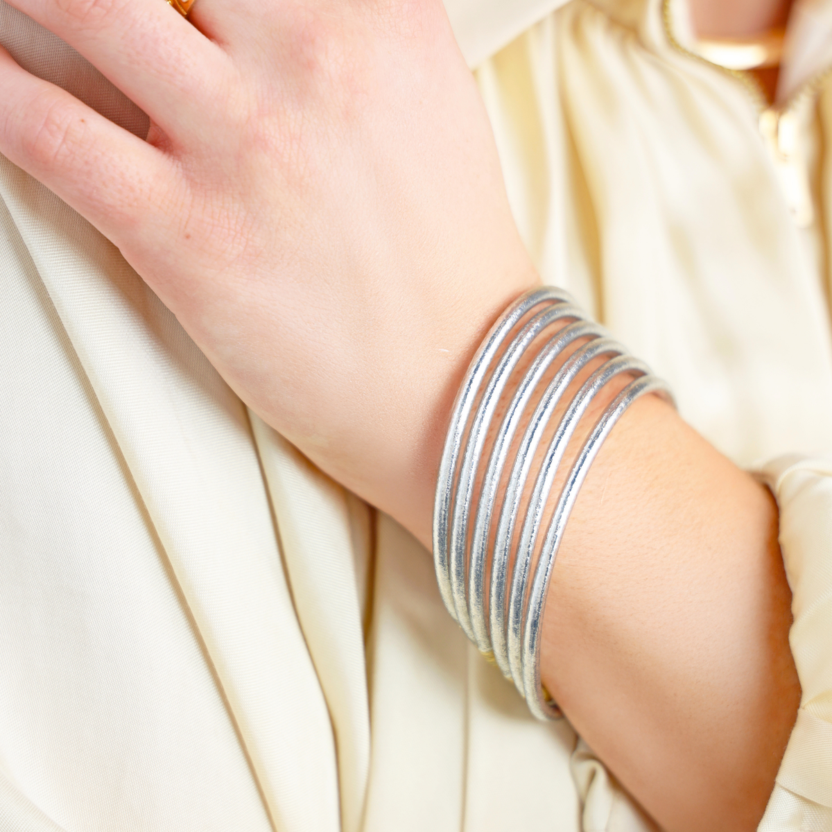 A close-up of a person's wrist wearing a stack of thin silver bangles over a cream-colored satin blouse.