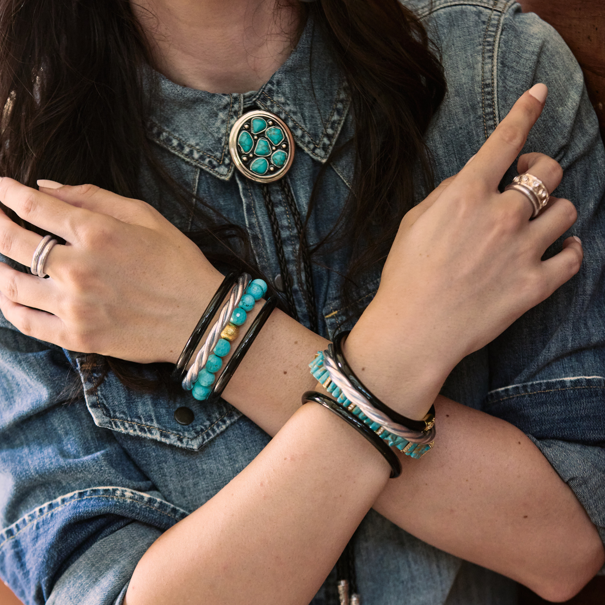 A close-up of a person wearing multiple turquoise and silver bracelets, a matching turquoise necklace, and silver rings, styled with a denim shirt.
