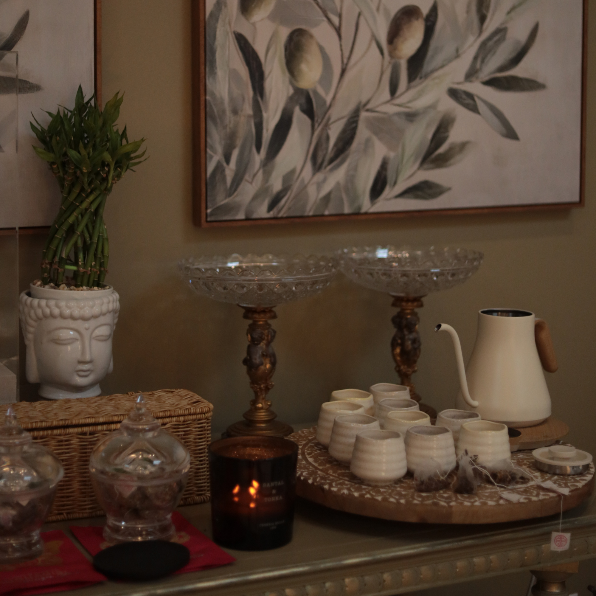 A styled interior tabletop display featuring a white ceramic Buddha head planter with bamboo, clear glass apothecary jars, two ornate brass candlesticks, a small amber candle, a white ceramic pitcher, and a tray of small candles in a warm, home decor setting.