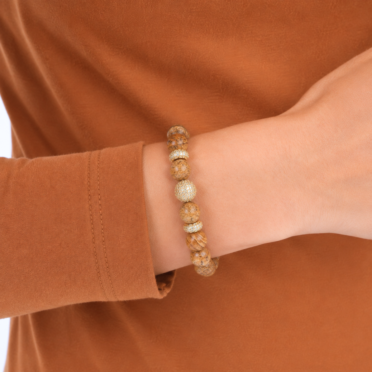 A close-up of a person's wrist wearing a beaded bracelet made of natural tan and brown wood beads with a textured silver-toned accent bead, set against a warm rust-brown long-sleeve shirt.