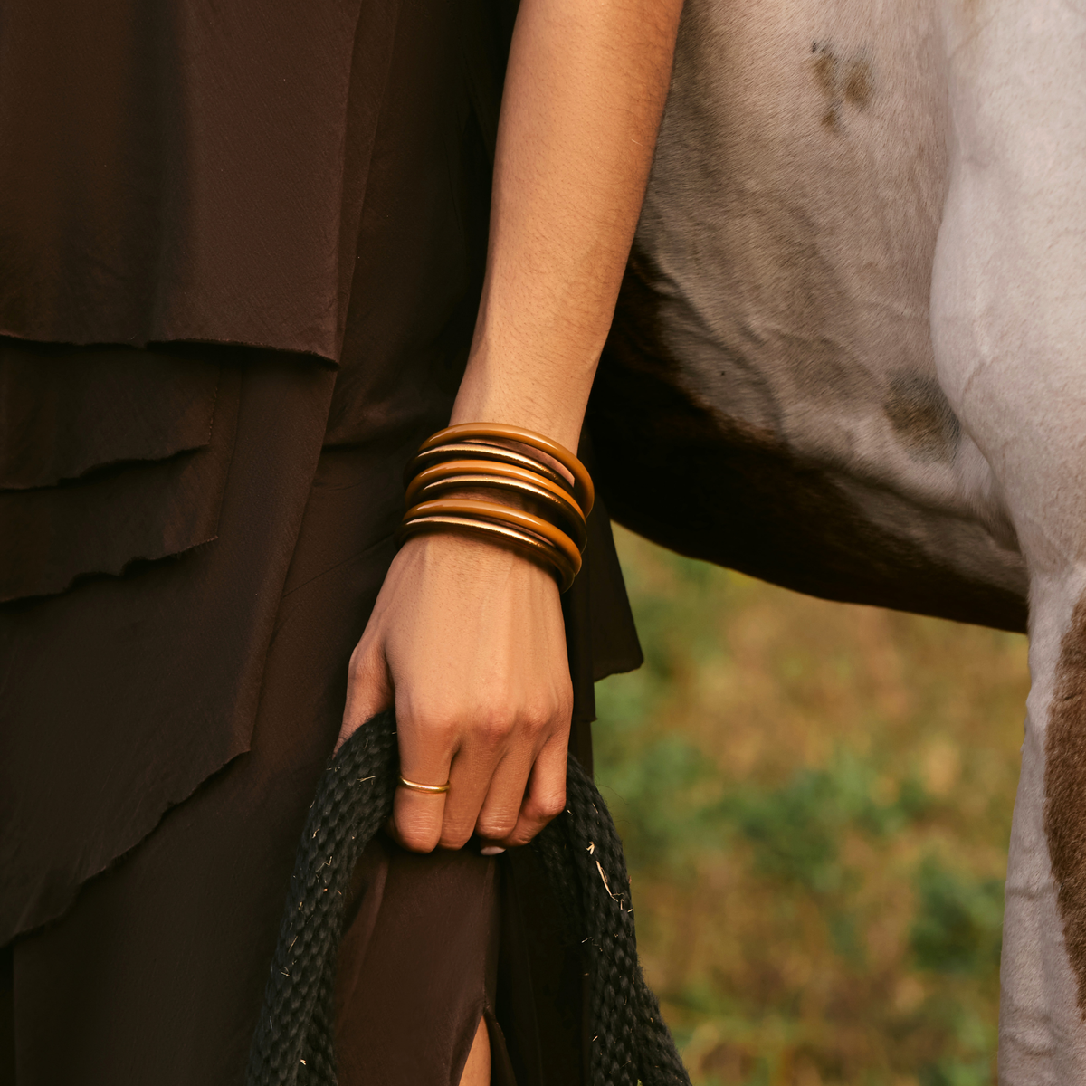 A close-up lifestyle shot of a person wearing a stack of bronze and brown bangles on their wrist, standing next to a horse outdoors.