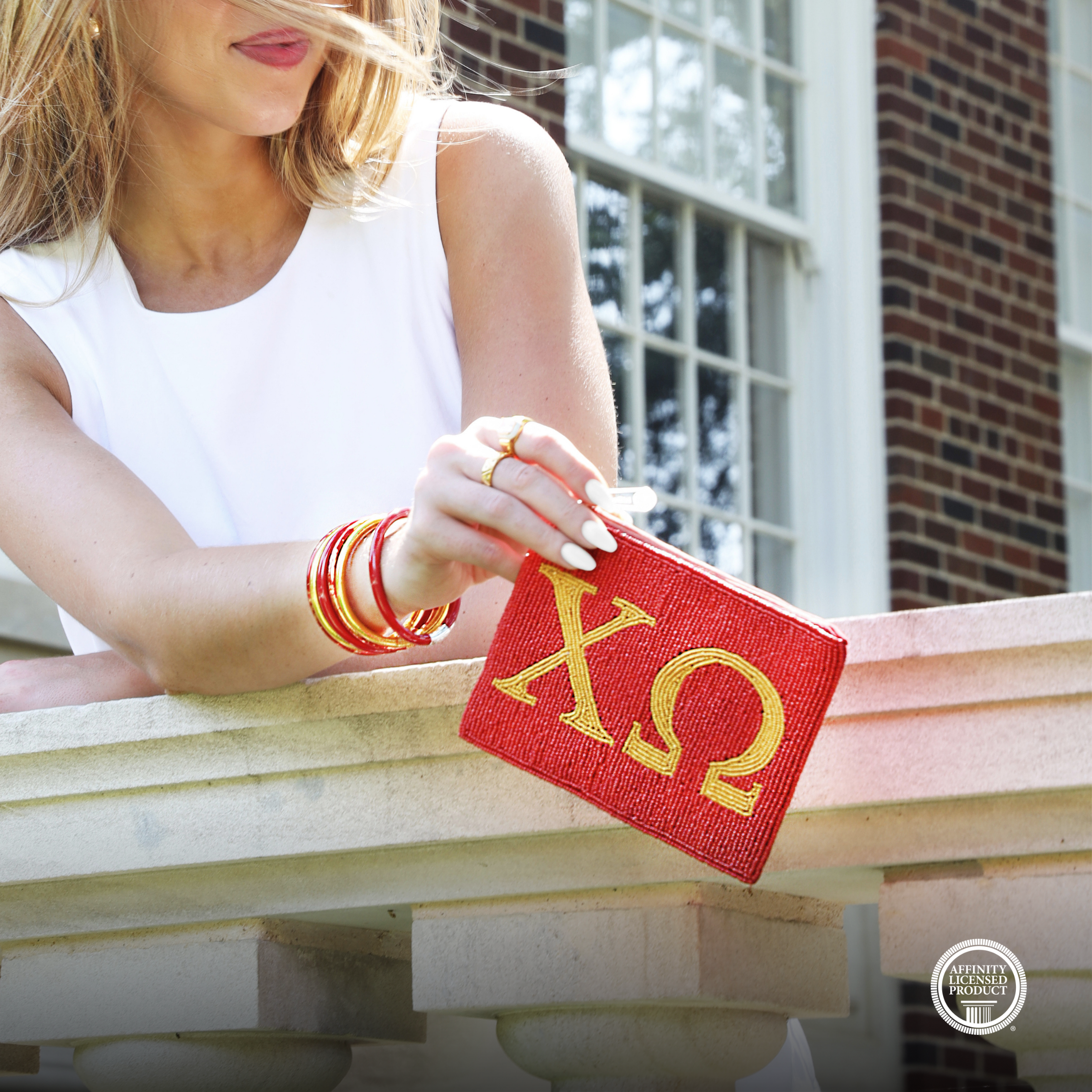 A woman in a white sleeveless top holds a red clutch bag with gold Greek letters (Chi Omega) in front of a brick building window. She is wearing red and gold bangles.